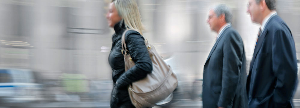 Woman in black jacket carrying a large beige handbag walking outdoors.